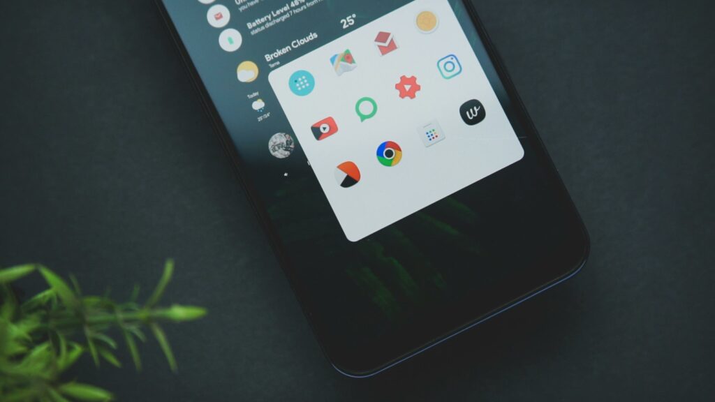 Close-up of a smart phone displaying various app icons on a dark background with greenery.